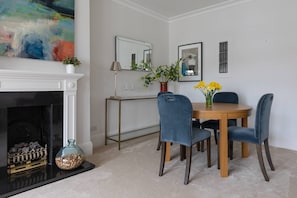 Ein Essbereich mit einem runden Holztisch, blau gepolsterten Stühlen, einer Vase mit gelben Blumen, einem Kamin mit weißem Sims und einem großen abstrakten Gemälde darüber.