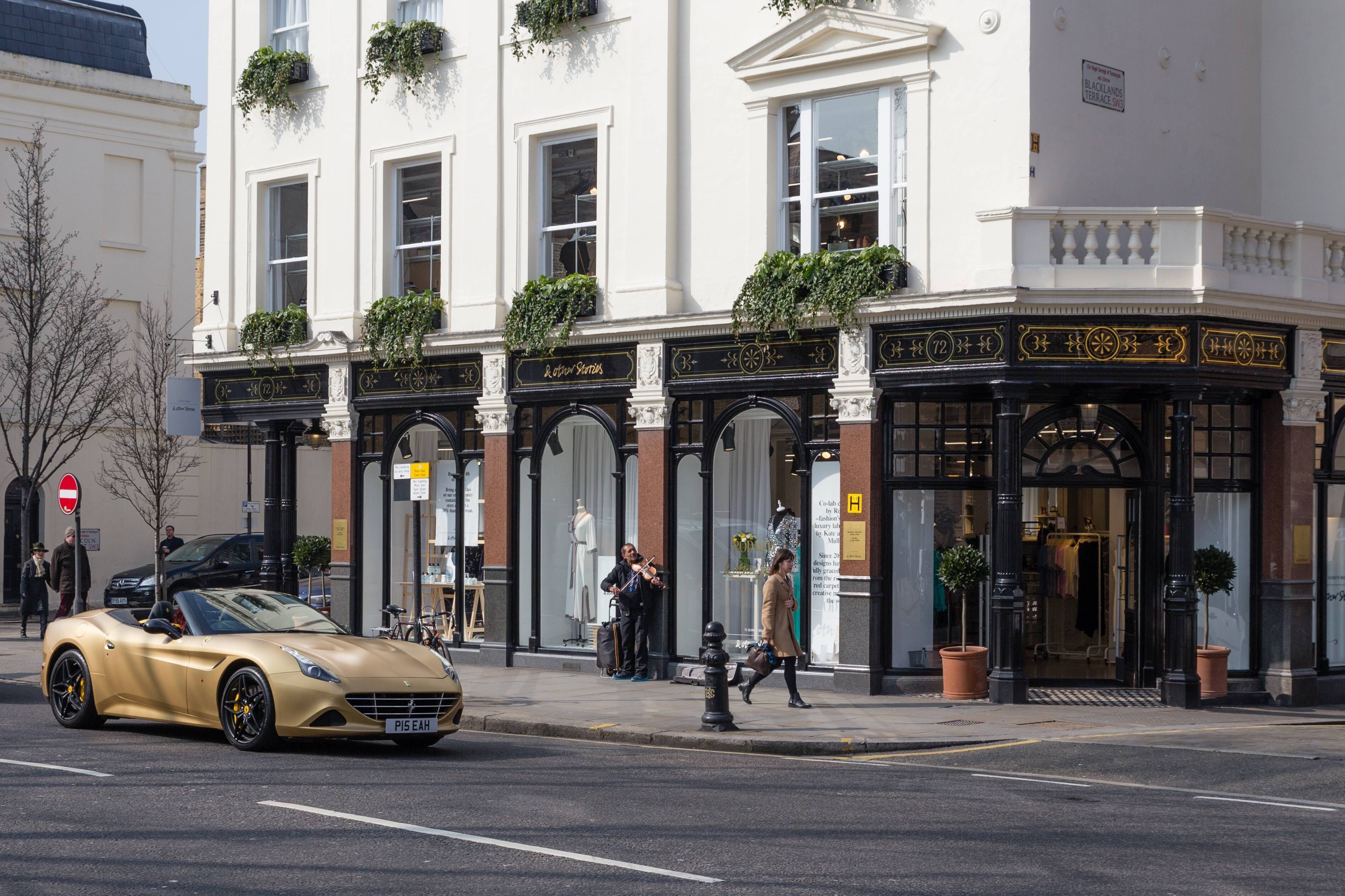 Une vue de rue avec un bâtiment doté d’un auvent noir, une enseigne indiquant « Aston Station », et une voiture décapotable dorée garée dans la rue.