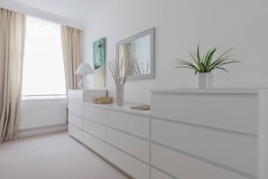 A white room with a large window, a white dresser, a lamp, a framed picture, and a potted plant. - Bathurst Mews III - Two Bedroom Apartment, Sleeps 4 (London)