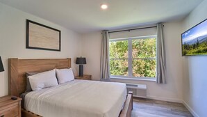 A bedroom with a wooden bed, a nightstand, a lamp, a window with curtains, and a framed picture on the wall. - The Ruby 311 (Pigeon Forge)