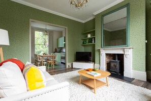 Ein Wohnzimmer mit einer Couch, einem Couchtisch, einem Kamin, einem Fernseher und einer durch einen offenen Durchgang einsehbarer Küche.