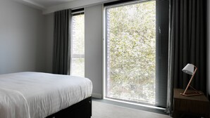 A bedroom with a bed, a nightstand, a lamp, and large windows with blinds. - Compton Street - Two Bedroom Apartment, Sleeps 4 (London)