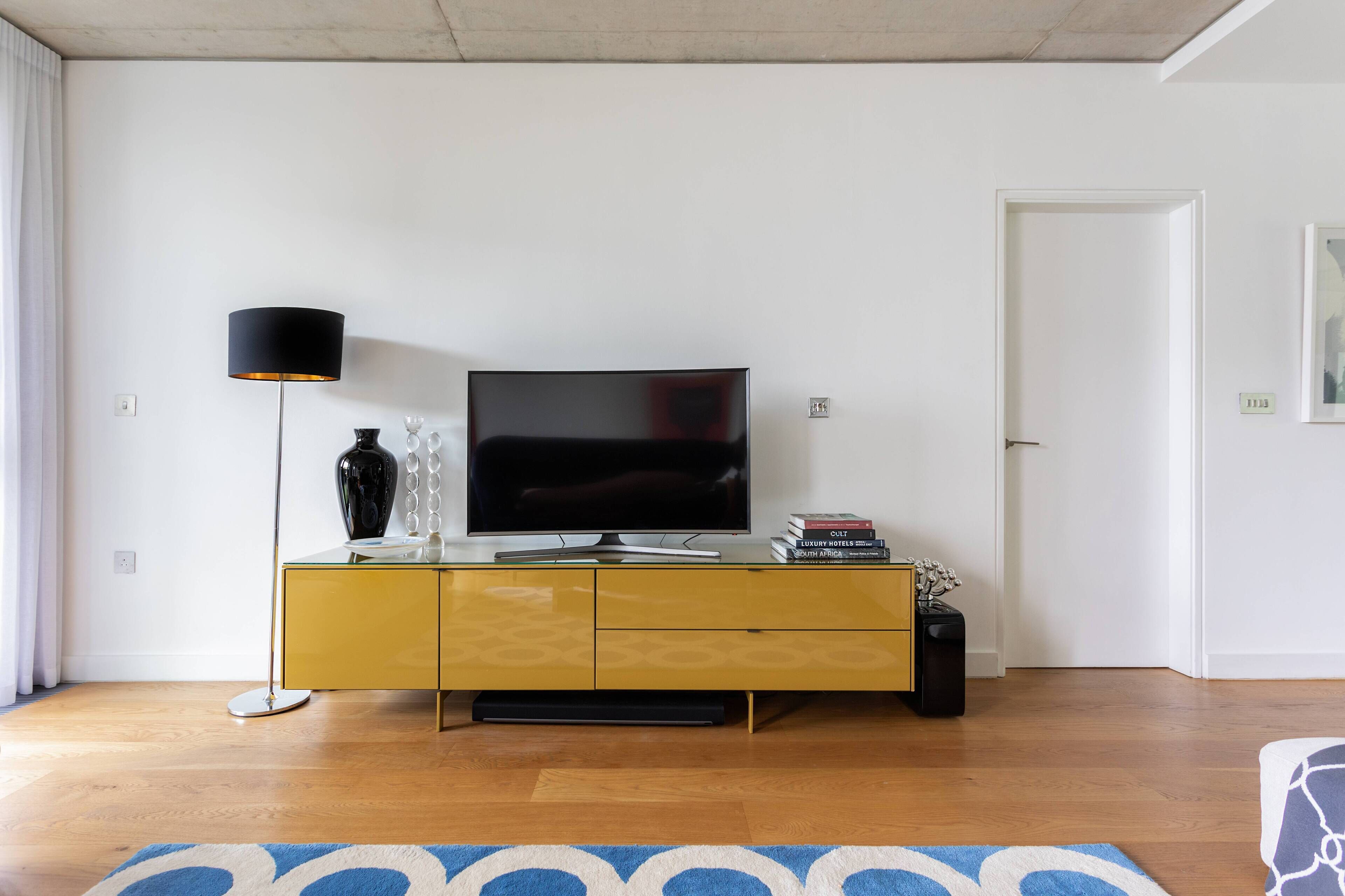 Un soggiorno moderno con televisore a schermo piatto su un mobiletto giallo, una lampada da terra e un tappeto decorato.