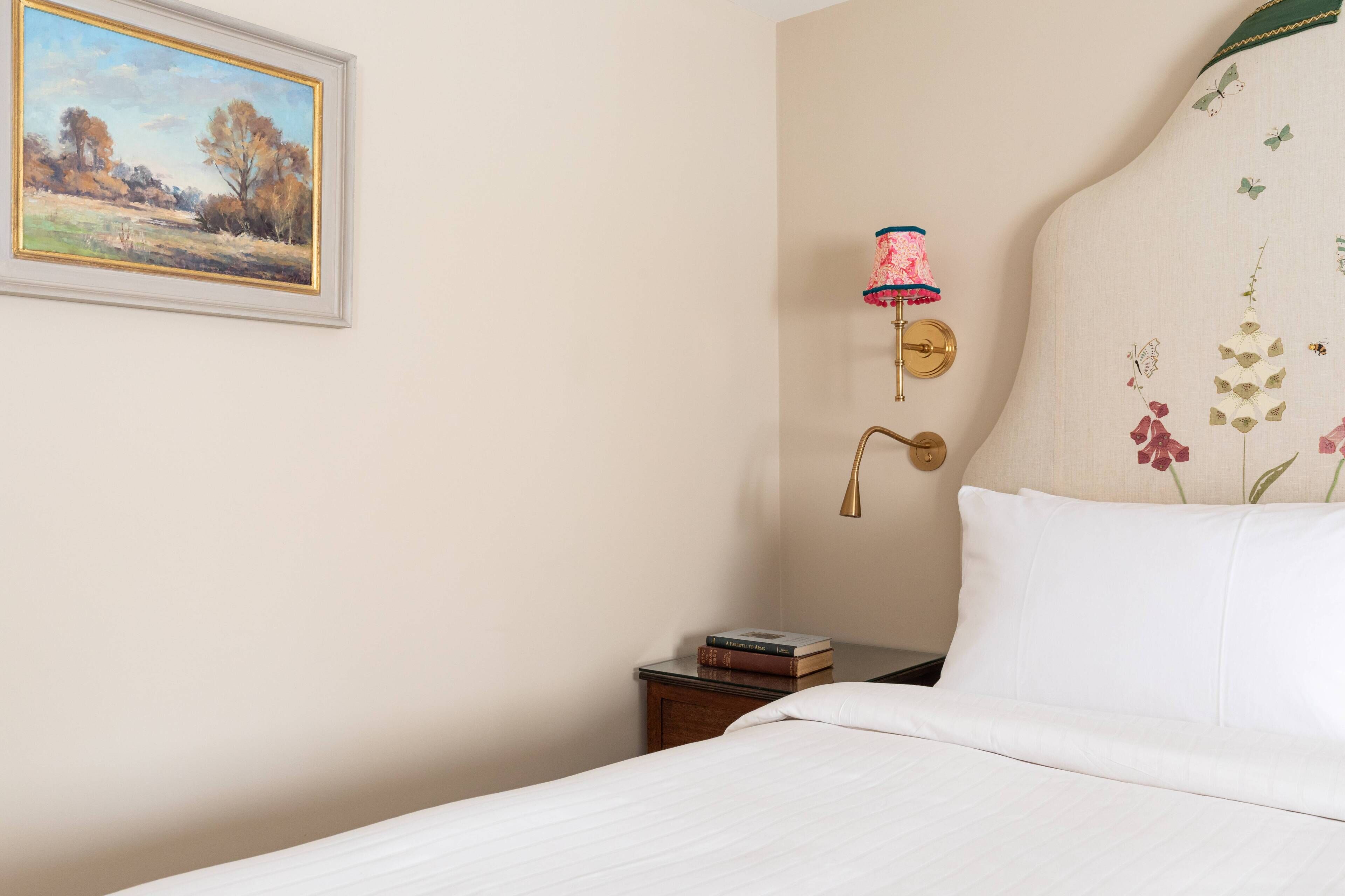 Una camera da letto con un letto, un comodino con dei libri, una lampada a parete e un quadro incorniciato.