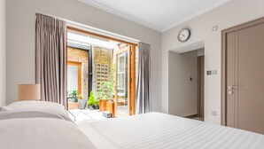 A hotel room with a bed, a lamp, a clock, and an open door leading to a balcony with plants. - Red Lion Square - Two Bedroom Apartment, Sleeps 4 (London)
