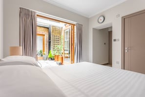 A hotel room with a bed, a lamp, a clock, and an open door leading to a balcony with plants. - Red Lion Square - Two Bedroom Apartment, Sleeps 4 (London)