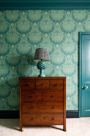 Eine hölzerne Kommode mit Schubladen und eine Lampe mit strukturierter Lampenschirm vor teal-farbenem Wandpapier mit Blättermuster.