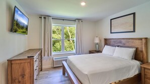 Un dormitorio con cama de madera, una mesita de noche, cómoda, ventana con cortinas y televisor montado en la pared.