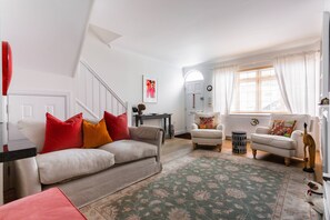 Ein Wohnzimmer mit einer Couch, zwei Sesseln, einem Couchtisch, einem Teppich, einer Treppe, einer Tür und einem Fenster mit Vorhängen.