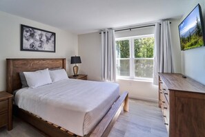 Un dormitorio con cama de madera, mesita de noche, lámpara, cómoda, ventana con cortinas e imagen enmarcadas en la pared.