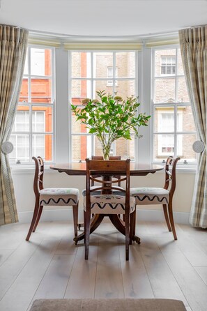 A dining area with a table and chairs, a potted plant, and large windows with curtains. - Tite Street VIII - One Bedroom Apartment, Sleeps 2 (London)