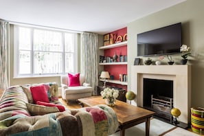 A living room with a sofa, armchair, coffee table, fireplace, television, and bookshelves. - Queens Gardens III - Two Bedroom Apartment, Sleeps 4 (London)