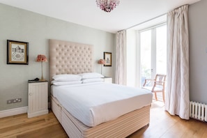 A bedroom with a large bed, a chest of drawers, a radiator, and a chair by the window. - Queens Gardens III - Two Bedroom Apartment, Sleeps 4 (London)
