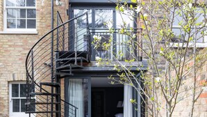 Ein zweigeschossiges Gebäude mit einer Wendeltreppe, die zu einem Balkon führt, einem kleinen Garten mit Pflanzen und einem sichtbaren Fenster.