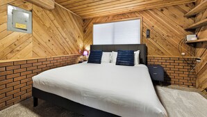 A bedroom with a wooden ceiling, a brick accent wall, a bed with white bedding, two dark pillows, a nightstand, and a window with blinds. - Rad Pad - Arcades, Seasonal Pool (Boise)