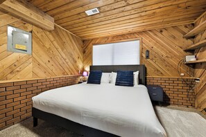 Ein Schlafzimmer mit Holzbalkendecke, einer Ziegelfassade, einem Bett mit weißer Bettwäsche, zwei dunklen Kissen, einem Nachttisch und einem Fenster mit Jalousien.