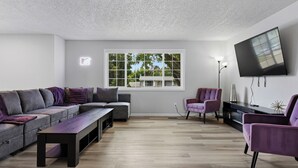 A modern living room with a sectional sofa, a bench, a flat-screen TV mounted on the wall, and a window with a view of trees. - Rad Pad - Arcades, Seasonal Pool (Boise)