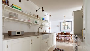 Una cocina con armarios blancos, una encimera con una radio, estanterías con platos y libros, un comedor con sillas y una mesa, y una ventana con vistas.