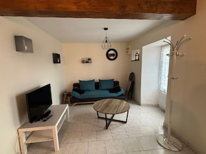 A living room with a sofa, a coffee table, a TV on a stand, a hanging light fixture, and a door with a coat rack. - House near the landing beaches Sainte Mere Eglise/Carentan (Boutteville)