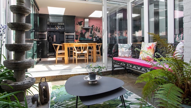 Eine Terrasse mit einer Steinskulptur, einem Esstisch mit Stühlen, einer Couch mit Kissen und einer Topfpflanze.