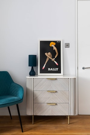 A framed artwork titled 'BALLY' on a white console with drawers, a teal chair, and a dark blue lamp. - St Stephen's Gardens VIII (London)