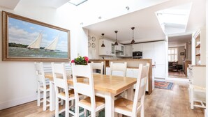Un comedor con una mesa de madera y sillas blancas, un cuadro de veleros y una cocina al fondo.
