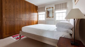 Un dormitorio con un armario de madera, una cama con ropa de cama blanca, una lámpara de noche y una ventana con persiana a rayas.