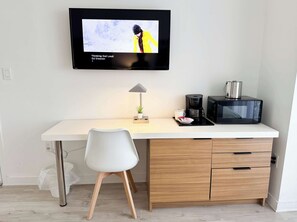 A modern kitchen with a flat-screen TV mounted above a white countertop, a coffee maker, a microwave, and a white chair. - Gorgeous 2BR - Beachwalk Intracoastal View (Hallandale Beach)