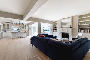 Ein großzügiger Wohnbereich mit einem dunkelblauen Sofa, einem weißen Kamin und einer Küche mit weißen Schränken und einer Bar.