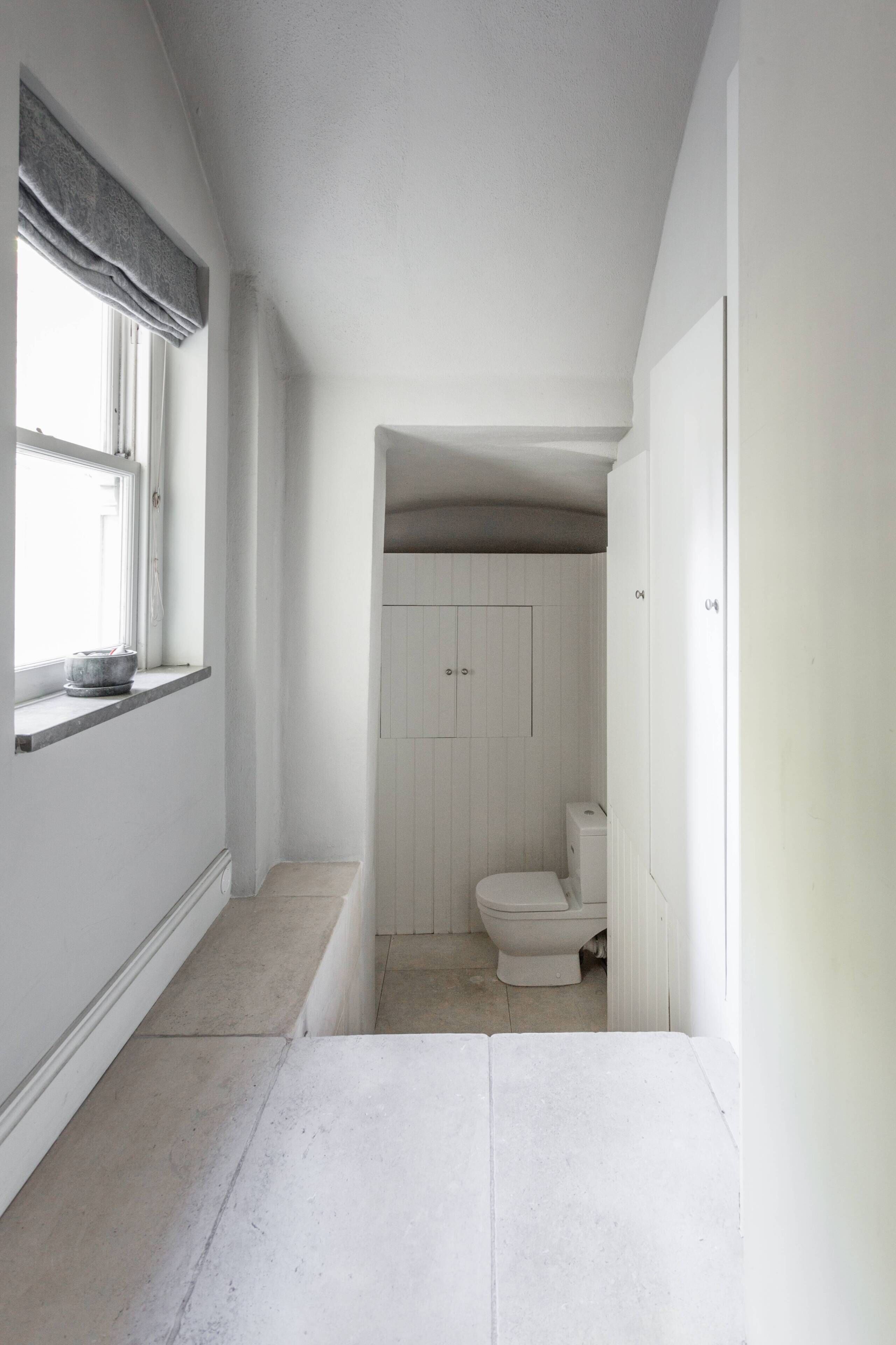 En smal hall som leder till ett badrum med en toalett och ett fönster med en gardin.