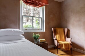 Ein Schlafzimmer mit einem Bett, einem Sessel, einem Fenster und einem Nachttisch mit Büchern und einer Vase.