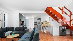 Ein Wohnzimmer mit Sofa, Couchtisch und Kamin. Es gibt eine Küchenzeile mit Esstisch und Stühlen, sowie einen Waschbereich mit Waschmaschine. Eine Holztreppe ist zu sehen.