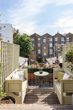 Un patio con una terraza de madera, dos sofás color beige, una mesita redonda y una planta en maceta.
