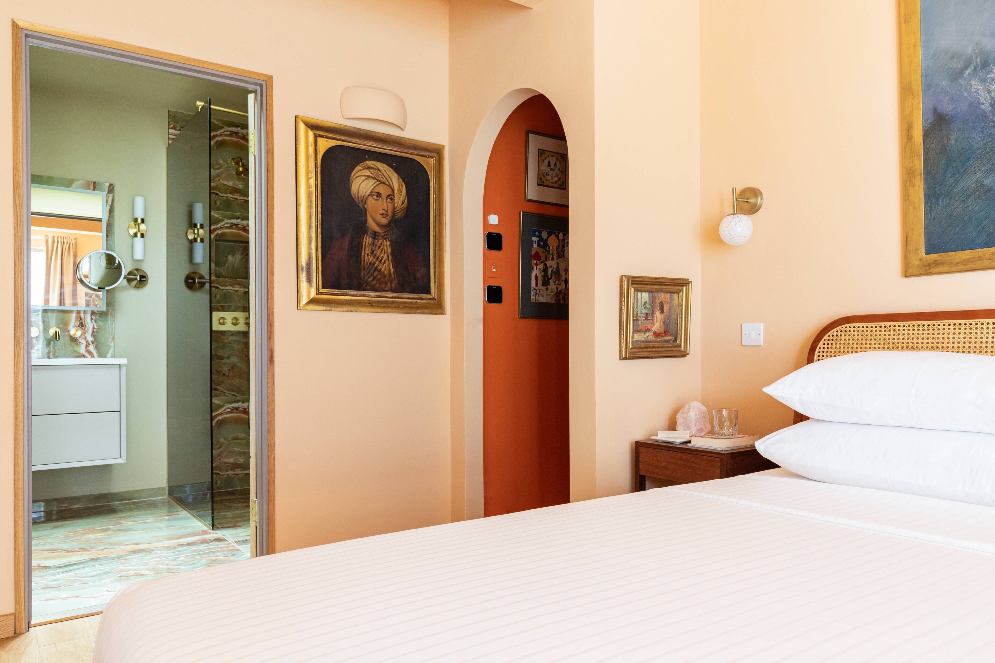 Een hotelkamer met een bed, een nachtkastje, een spiegel, een lijst met een portret en een deur die naar een andere kamer leidt.