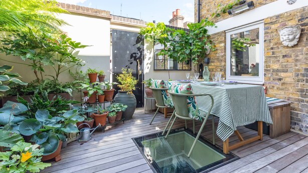 Eine Terrasse mit Tisch und Stühlen, umgeben von Blumentöpfen und einer Ziegelwand.