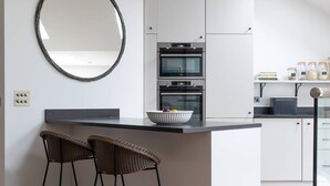 A modern kitchen with a central island, two bar stools, a round mirror, and built-in ovens. - Granard Road - Four Bedroom House, Sleeps 8 (London)