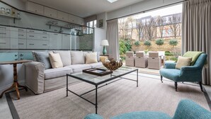 A modern living room with a glass coffee table, sofa, and armchair, a kitchen with cabinets and a window, and a view of the outside. - Granard Road - Four Bedroom House, Sleeps 8 (London)