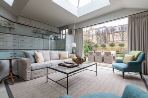 Ein modernes Wohnzimmer mit Glastisch, Sofa und Sessel, eine Küche mit Schränken und einem Fenster, und ein Blick ins Freie.