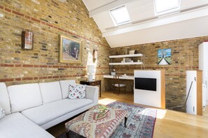Ein Loft-Zimmer mit Backsteinwand, einem weißen Sofa, einem gemusterten Hocker, einem Schreibtisch und einem Fernseher.