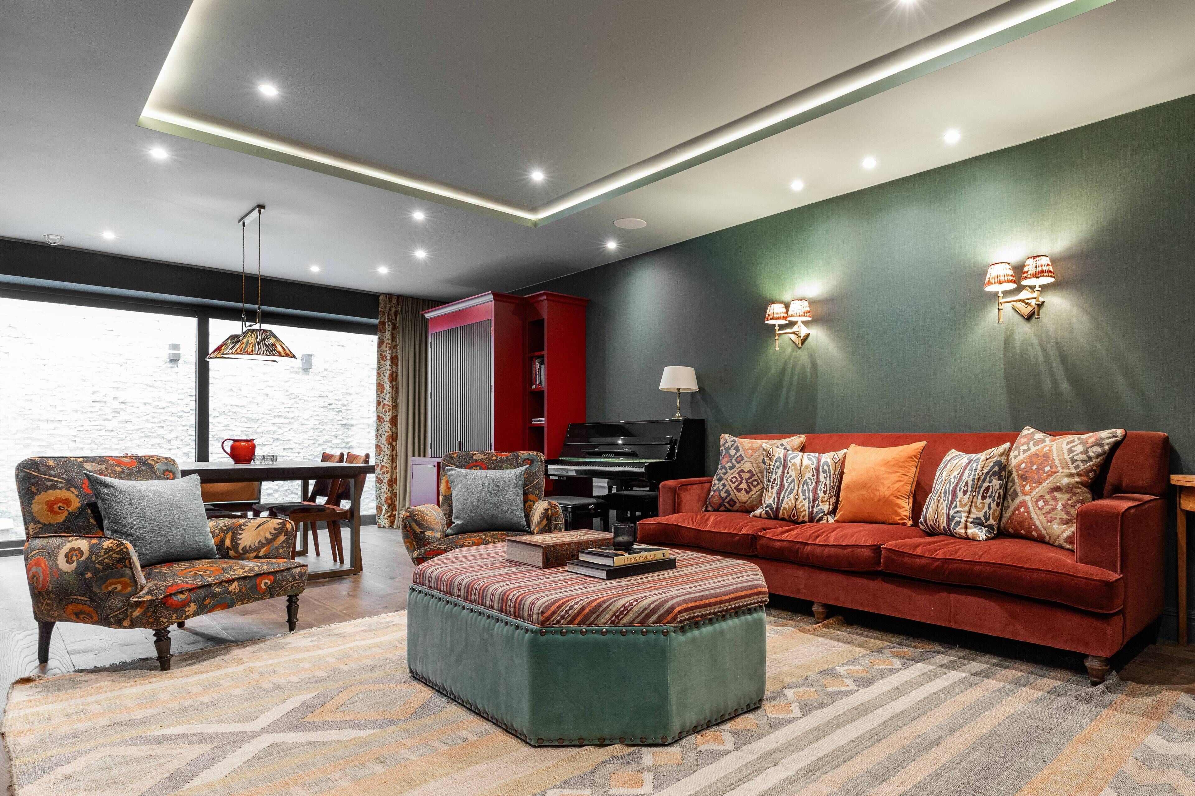 A living room with a red sofa, a patterned armchair, a green ottoman, a piano, and a view through a large window.