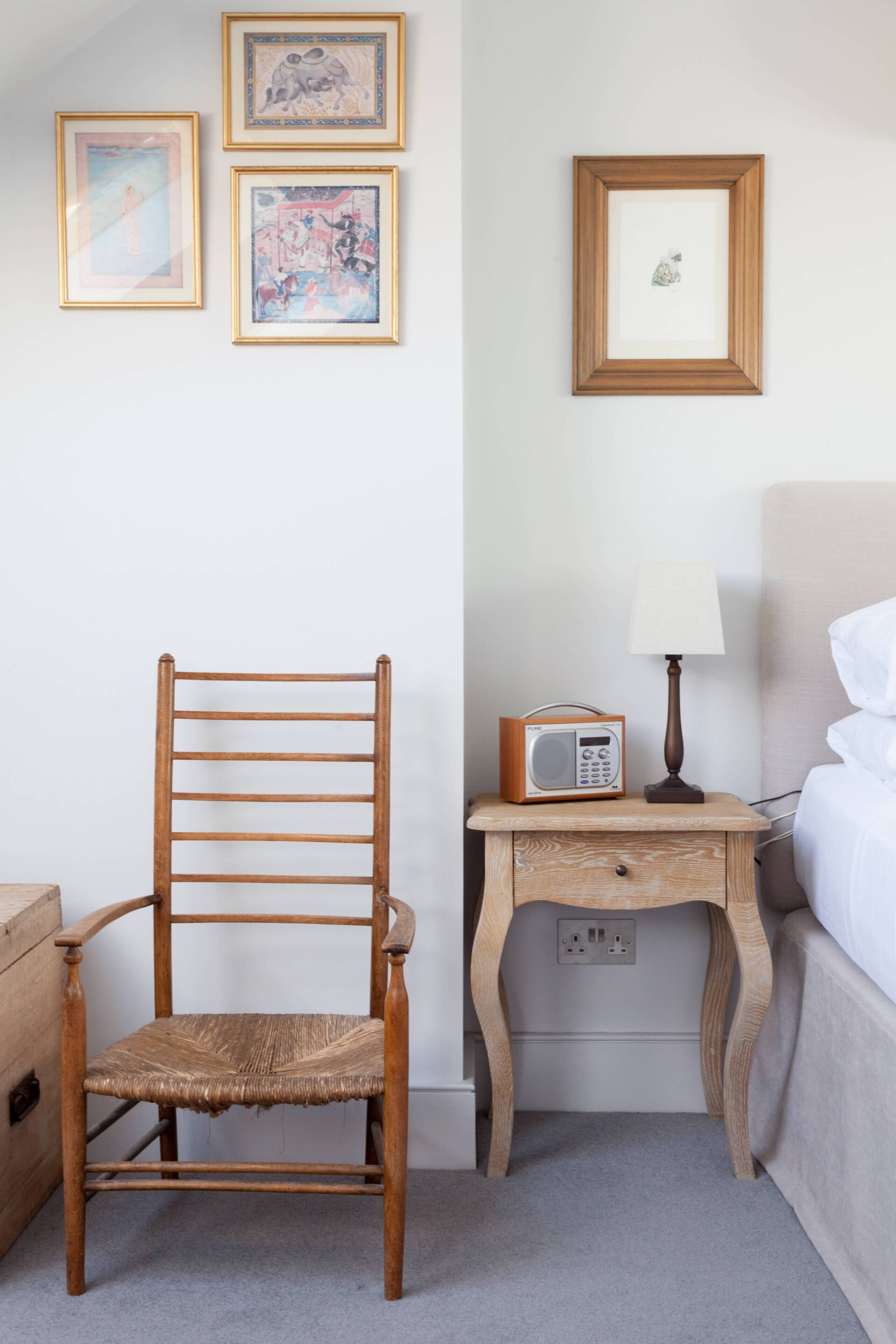A wooden chair, a bedside table with a radio, and framed pictures on the wall.