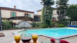 Une scène au bord de la piscine avec une table, deux verres contenant une boisson, un livre et un sac rouge.