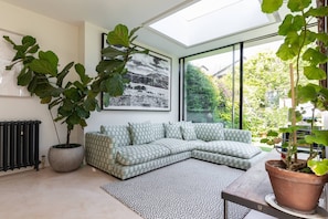Ein modernes Wohnzimmer mit einem Ecksofa, großen Kübelpflanzen, einem gerahmten Kunstwerk und einem Oberlicht.