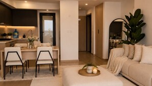 Ein modernes Wohnzimmer mit einer Couch, Hocker und Sesseln, eine Küchenzeile mit Schränken und einer Arbeitsplatte sowie ein Gang zu einem weiteren Raum.