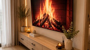 Ein modernes Wohnzimmer mit einem großen Flachbildfernseher an der Wand, einem Holztischchen mit einer Vase und Kerzen sowie einer Topfpflanze.