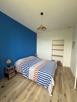 Une chambre à coucher avec un lit, une table de chevet, une lampe, un luminaire suspendu, un plancher en bois et un mur blanc avec une étagère.