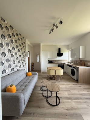 Un salon moderne avec un canapé gris, deux tables d’appoint en bois, un mur à motifs et une cuisine dotée de meubles blancs et d’un lave-linge.
