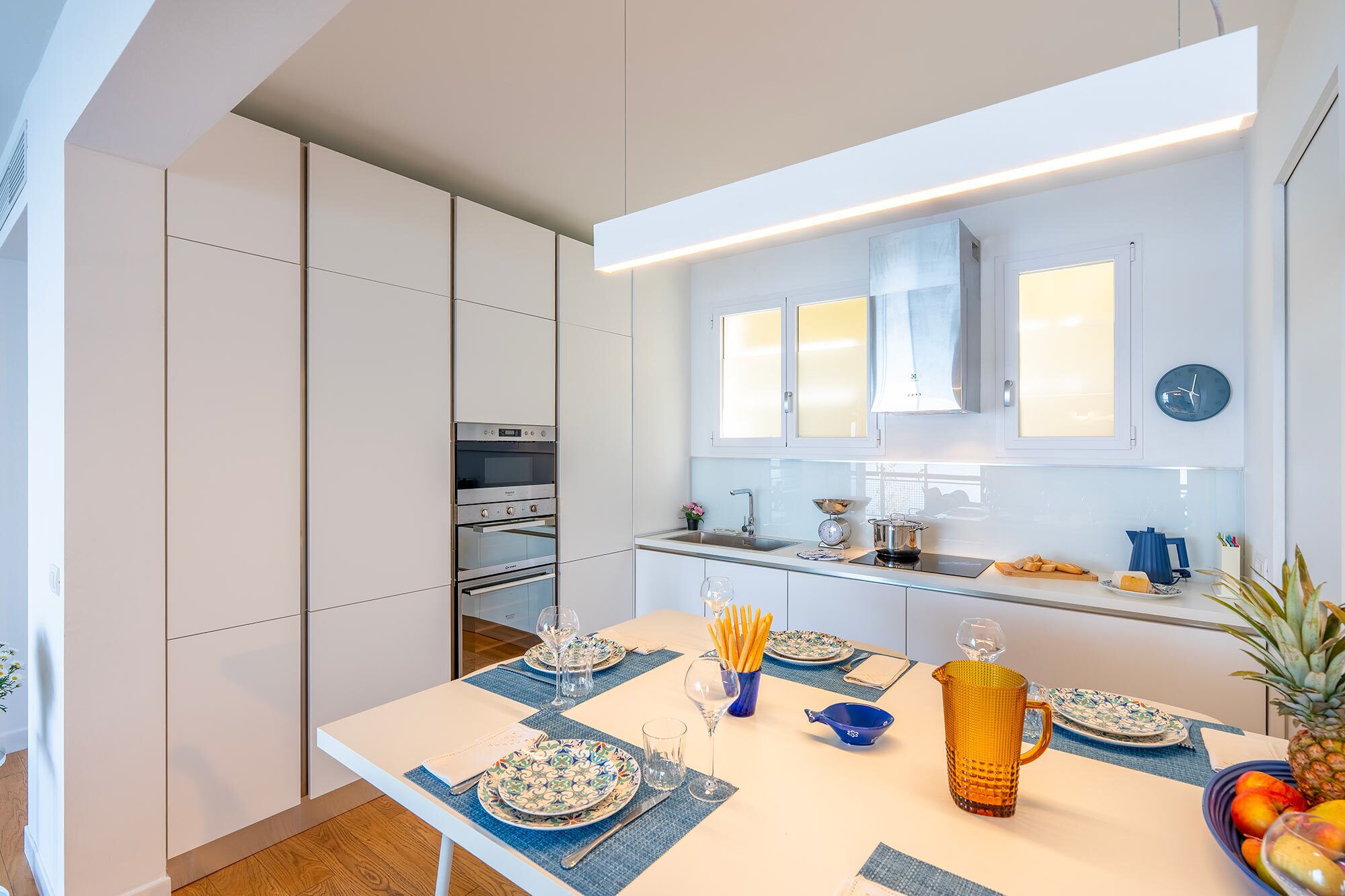 Una cucina moderna con elettrodomestici integrati, un tavolo da pranzo apparecchiato con piatti e bicchieri, e una ciotola di frutta sul piano di lavoro.