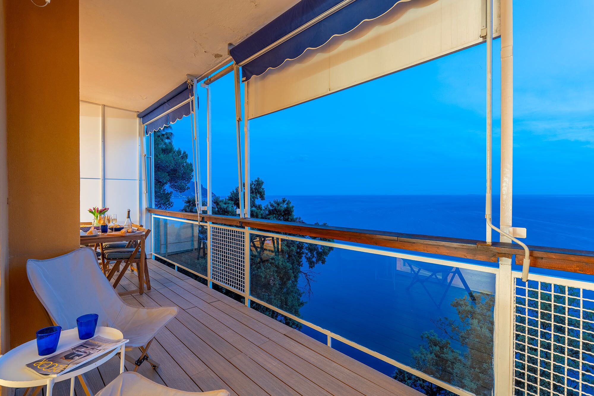 Uma varanda com vista para o mar, uma mesa com cadeiras e uma janela com vista para árvores e o mar.