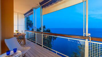 Un balcone con vista sul mare, un tavolo con sedie e una finestra che si affaccia su alberi e il mare.